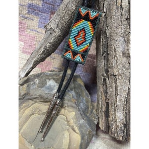 May include: A bolo tie with a rectangular beaded pendant. The pendant has a turquoise, orange, and yellow geometric design on a black background. The tie has two black braided cords and silver-coloured tips.