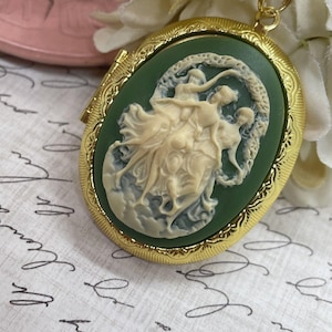 Puede incluir: Un medallón de oro con un camafeo verde que presenta una escultura en relieve blanca de tres mujeres bailando. El camafeo está engastado en un marco ovalado de oro con un borde decorativo.