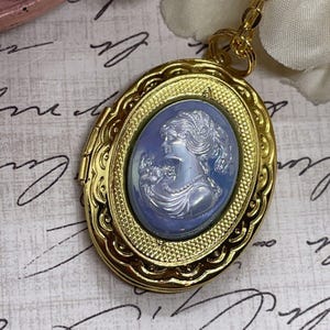 Könnte beinhalten: Ein goldenes Medaillon mit einem blauen Kamee, der das Profil einer Frau zeigt. Der Kamee ist in einen goldenen Rahmen mit einem dekorativen Rand gefasst.