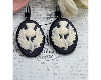 May include: Black oval earrings with a cream-colored thistle design. The thistle is surrounded by cream-colored leaves. The earrings have black lever-back closures. The earrings are set against a vintage paper background.