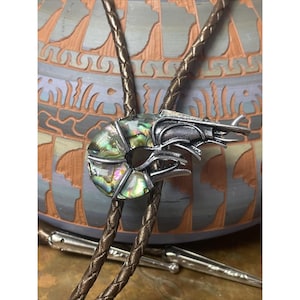 May include: A bolo tie featuring a silver-toned shrimp pendant with iridescent abalone shell accents. The shrimp is attached to a brown braided leather cord with silver tips. The background is a decorative ceramic vase.