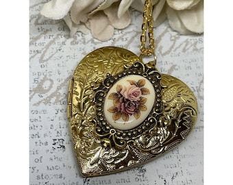May include: A gold-colored heart-shaped locket with an oval floral cameo. The locket has ornate detailing and hangs from a gold chain. The cameo features a pink and burgundy rose bouquet on a cream background.