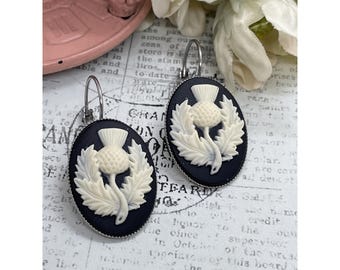 May include: A pair of oval-shaped earrings featuring a white thistle design on a black background. The earrings have a silver-colored hook closure. The thistle is surrounded by leaves. The earrings are set against a vintage paper background.