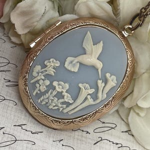 May include: Oval gold-tone locket with a light blue cameo featuring a white hummingbird and floral design. The locket has a detailed border and a hinged opening. A delicate chain is attached.