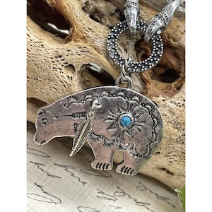 May include: Silver-toned bear pendant necklace with turquoise stone and feather accents. The bear is intricately detailed with etched designs and an arrow. The necklace features a large, decorative ring and clasp.