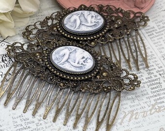 May include: Two antique bronze hair combs. Each comb features an oval cameo with a white relief design and is surrounded by an ornate filigree frame. The combs have multiple tines and are designed for securing hair.