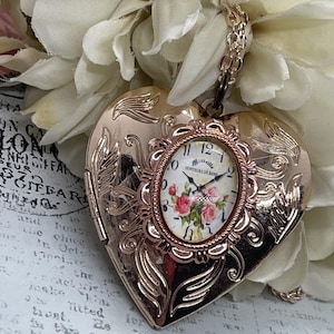 Peut inclure: Un collier avec un médaillon en forme de cœur en or rose et un cadran ovale. Le cadran de l'horloge présente un motif floral avec des roses roses et le texte "Sentiers de Rose". Le médaillon est orné de gravures florales.