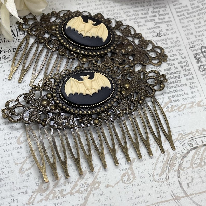 Op de afbeelding: Twee antieke bronzen haar kammen. Elke kam heeft een ovale cameo met een cr&egrave;me vleermuis op een zwarte achtergrond. De kammen hebben ingewikkelde filigraan details, ideaal voor Halloween of gotische stijlen.