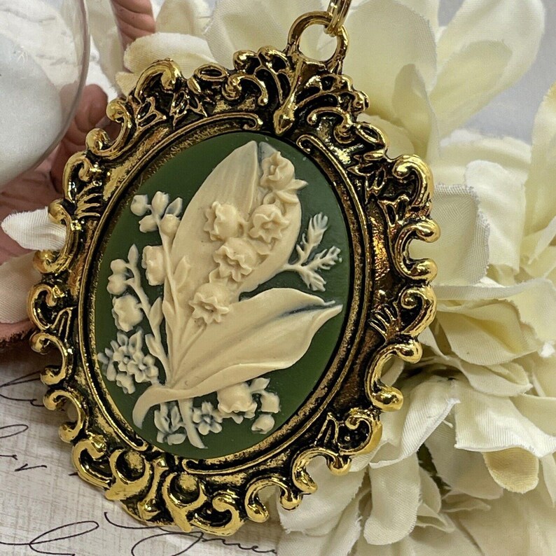 May include: A gold-tone cameo pendant featuring a white lily of the valley flower design on a green background. The pendant is set in an ornate oval frame.