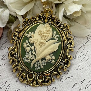 May include: A gold-tone cameo pendant featuring a white lily of the valley flower design on a green background. The pendant is set in an ornate oval frame.