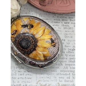 Puede incluir: Colgante ovalado plateado con un diseño de girasol y abejas. El interior del relicario muestra un girasol amarillo con abejas negras y amarillas. El fondo tiene escritura vintage. El colgante está en una cadena.
