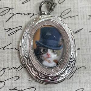 May include: Silver oval locket necklace with a portrait of a cat wearing a top hat and monocle.