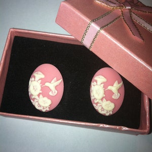Peut inclure: Boucles d'oreilles ovales roses avec un motif de colibri et de fleurs, présentées dans un coffret cadeau rose assorti avec un ruban. Les boucles d'oreilles ont un motif de colibri et de fleurs blanches sur fond rose.