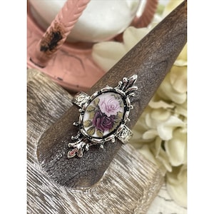 May include: A silver ring featuring an oval cameo with a floral design of pink and purple roses. The cameo is set in a silver frame with ornate details.
