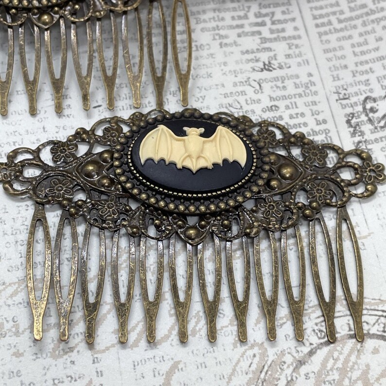 Op de afbeelding: Antieke bronzen haar kam met een decoratief filigraan ontwerp. De kam heeft een zwarte ovale cameo met een cr&egrave;mekleurige vleermuis. De tanden van de kam zijn gelijkmatig verdeeld, wat een vintage, gotische uitstraling geeft.