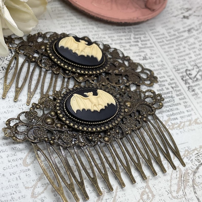 Op de afbeelding: Twee antieke bronzen haar kammen. Elke kam heeft een ovale camee met een cr&egrave;mekleurige vleermuis op een zwarte achtergrond, gezet in een sierlijk filigraan frame. De kammen hebben meerdere tanden om in het haar te bevestigen.