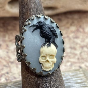 Op de afbeelding: Antieke ring met een zwarte raaf die bovenop een crèmekleurige schedel zit. De raaf staat tegen een lichtblauwe ovale achtergrond, omlijst door een decoratief metaal in bronskleur. Een uniek gothic-thema accessoire.