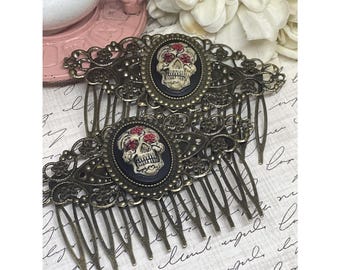 May include: Two antique bronze-toned hair combs. Each comb features an oval centerpiece with a black background and a white skull with red floral accents. The combs have intricate filigree designs and multiple teeth.