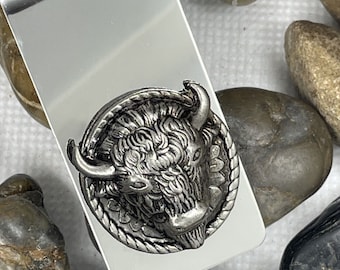 May include: A silver-colored money clip with a detailed, antique-style buffalo head emblem. The buffalo head is in a circular frame with a rope-like border. The clip is rectangular and has a shiny, reflective surface.