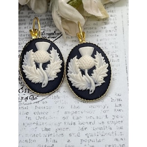 May include: A pair of oval earrings with a thistle design. The earrings have a black background with a cream-coloured thistle and gold-coloured hooks. The thistle is surrounded by leaves.