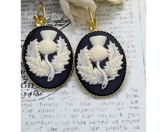 May include: A pair of oval earrings featuring a thistle design. The earrings have a black background with a cream-colored thistle and gold-colored hooks. The thistle is surrounded by leaves.
