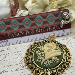 May include: A gold-tone pendant necklace featuring a cameo of white lily of the valley flowers on a green background. The cameo is set in a decorative gold frame. The necklace is displayed on a ruler with the text "FANCY FOX BOUTIQUE" printed on it.