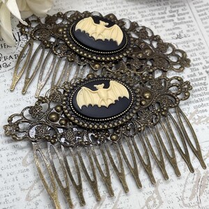 Op de afbeelding: Twee antieke bronzen haar kammen. Elke kam heeft een zwarte ovale cameo met een cr&egrave;me vleermuis ontwerp. De kammen hebben ingewikkelde filigraan details en lange, dunne tanden. Een gotisch of Halloween-thema accessoire.