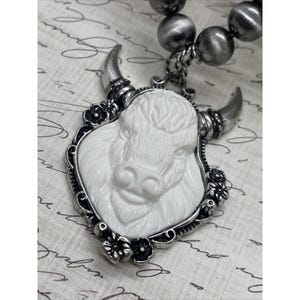 May include: A white buffalo pendant necklace with a silver-tone frame and horns. The buffalo is carved in detail, and the frame is decorated with small flowers. The necklace has a strand of gray beads.