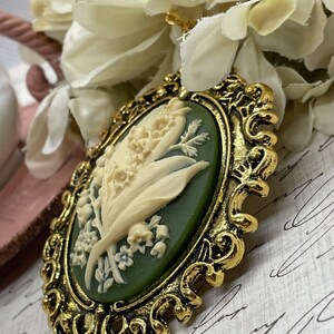 May include: A gold-tone cameo pendant featuring a white lily of the valley flower design on a green background.