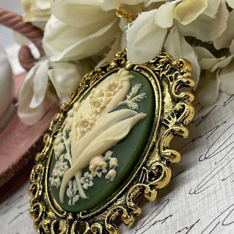 May include: A gold-tone cameo pendant featuring a white lily of the valley flower design on a green background.