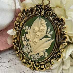 May include: A gold-tone cameo pendant featuring a white lily of the valley flower design on a green background. The pendant is set in an ornate oval frame.