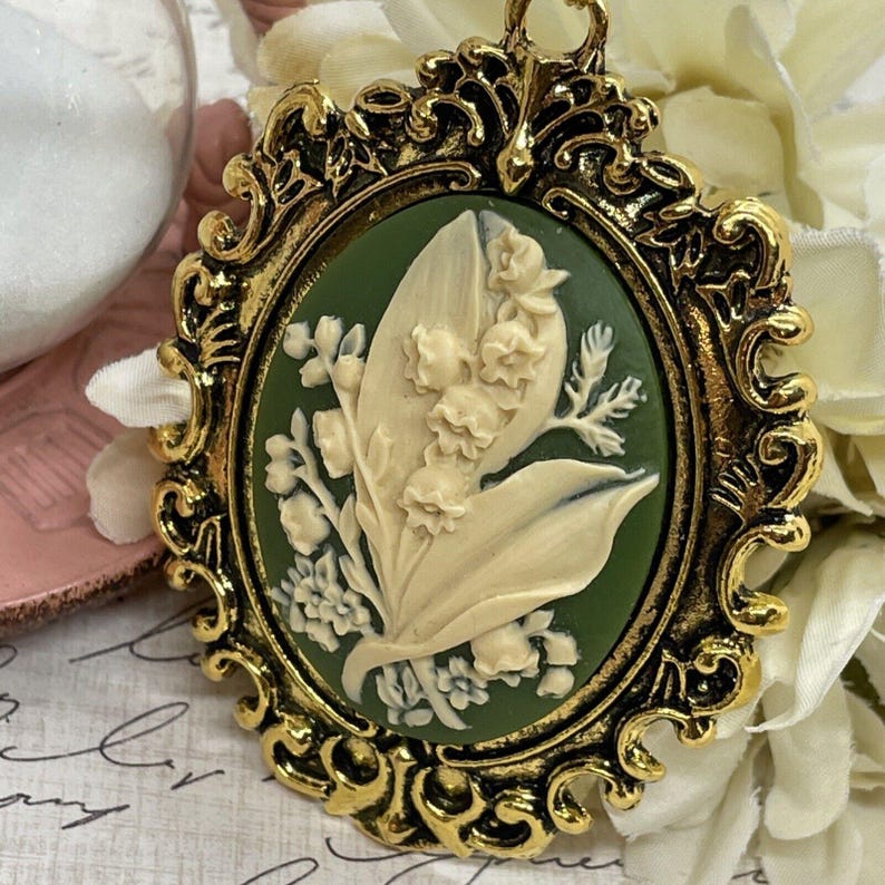 May include: A gold-tone cameo pendant featuring a white lily of the valley flower design on a green background. The pendant is set in an ornate oval frame.