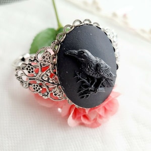 May include: A black cameo bracelet with a silver filigree design. The cameo features a black raven perched on a branch.