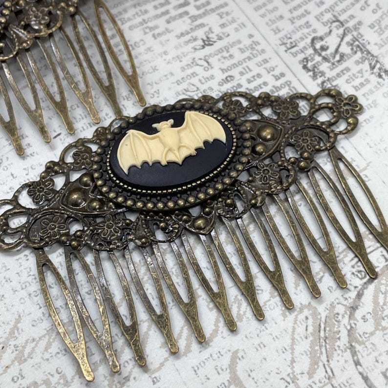 Op de afbeelding: Antieke bronzen haarspeld met een ovale zwarte camee met een cr&egrave;mekleurige vleermuis. De kam heeft ingewikkelde filigraan details en meerdere tanden. Een gotisch ge&iuml;nspireerd haaraccessoire.