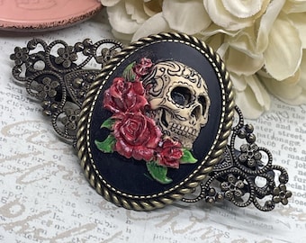 May include: An oval-shaped hair clip featuring a skull design with red roses. The skull is cream-colored with intricate detailing, set against a black background. The clip has a bronze-colored filigree frame.