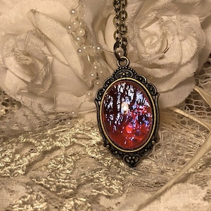 May include: A red and purple iridescent glass pendant necklace with an ornate bronze frame. The necklace is on a white lace background.