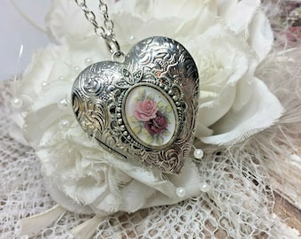 May include: Silver heart-shaped locket necklace with a floral design. The locket has a hinged opening and a small chain attached to the top. The locket is on a white background.