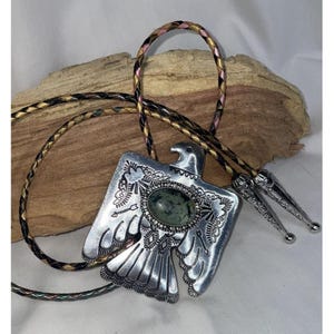 May include: A silver bolo tie featuring an eagle design with a green stone in the center. The bolo tie is on a braided leather cord with black, gold, and pink strands.