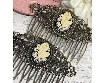 May include: Two antique bronze-toned hair combs. Each comb features an oval cameo with a cream-colored floral design set against a black background. The combs have intricate filigree detailing and long, thin teeth.