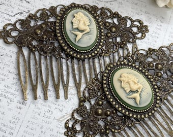 May include: Two antique bronze hair combs. Each comb features an oval cameo with a cream-coloured mermaid design set against a pale green background. The combs have intricate filigree detailing.