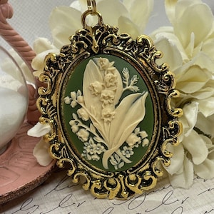 May include: A gold-tone cameo pendant featuring a white lily of the valley flower design on a green background. The pendant is in an oval frame with ornate details.