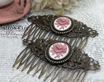 May include: Two antique bronze hair combs. Each comb features an oval cameo with a pink rose design on a white background. The combs have intricate filigree detailing and long, thin teeth.