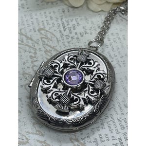 May include: An oval silver-toned metal locket pendant on a matching chain. The locket features an intricate raised design of four thistle motifs surrounding a central round, faceted light purple gemstone. The metal surface has a polished finish.