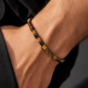 May include: A beaded bracelet featuring alternating square-shaped tiger eye beads in shades of golden brown and dark charcoal gray, arranged in a repeating pattern along a thin, flexible cord that encircles the wrist.