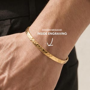 May include: A gold-toned cuff bracelet with a hammered texture and a width of 6mm. The bracelet features an open design and includes text on the image indicating an inside engraving option with a hidden message.