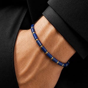Könnte beinhalten: Ein Armband aus zylindrischen, tiefblauen Lapislazuli-Perlen, die sich mit kleinen, polierten goldfarbenen Metallzwischenstücken abwechseln. Das Armband ist auf einer flexiblen Schnur aufgezogen und liegt auf einem schwarzen Stoffärmel.
