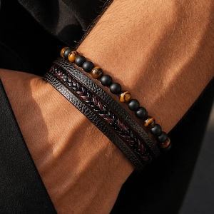 Puede incluir: Un conjunto de pulseras superpuestas que consta de una hilera de cuentas de piedra negra mate y ojo de tigre, y una segunda banda más ancha con un centro de cuero trenzado marrón oscuro flanqueado por bordes de cuero negro texturizado.