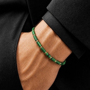 Pode incluir: Uma pulseira de contas cilíndricas de malaquita verde alternadas com pequenos espaçadores de metal dourado. A pulseira é usada no pulso, exibindo os padrões naturais de faixas da pedra verde e os detalhes metálicos.