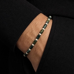May include: A beaded bracelet featuring alternating cylindrical dark green mottled stone beads and small, smooth off-white spacer beads. The bracelet is strung on a thin cord and displays a repeating pattern of the green and white components.