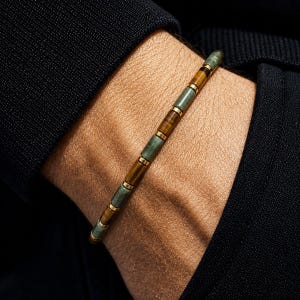 May include: A beaded bracelet featuring alternating cylindrical beads in shades of sage green and brown tiger eye, separated by small gold-toned metal spacers. The bracelet is worn on a wrist against a black fabric sleeve, showcasing a linear design.
